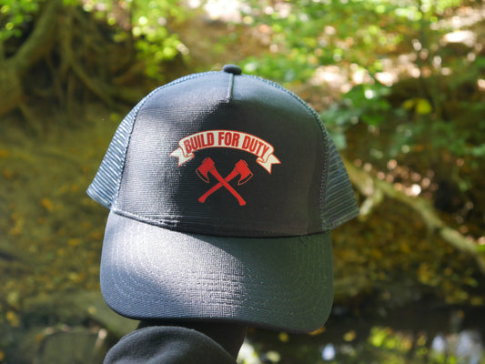 Dunkelblaue Mesh-Cap mit rotem „BUILD FOR DUTY“ Logo liegt im Waldlicht, Hintergrund unscharf mit Blättern und Natur
