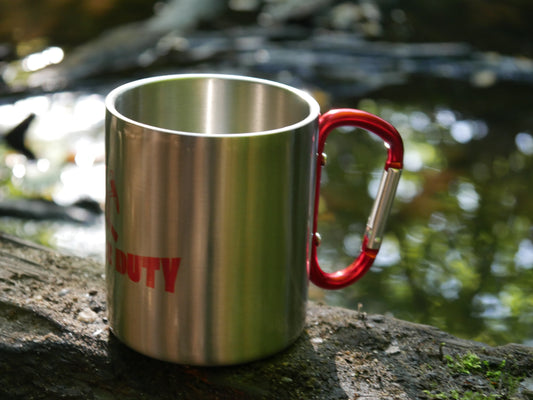Edelstahlbecher mit rotem „BUILD FOR DUTY“ Motiv steht auf einem moosigen Stein am Bachufer, mit unscharfem Wasser im Hintergrund.